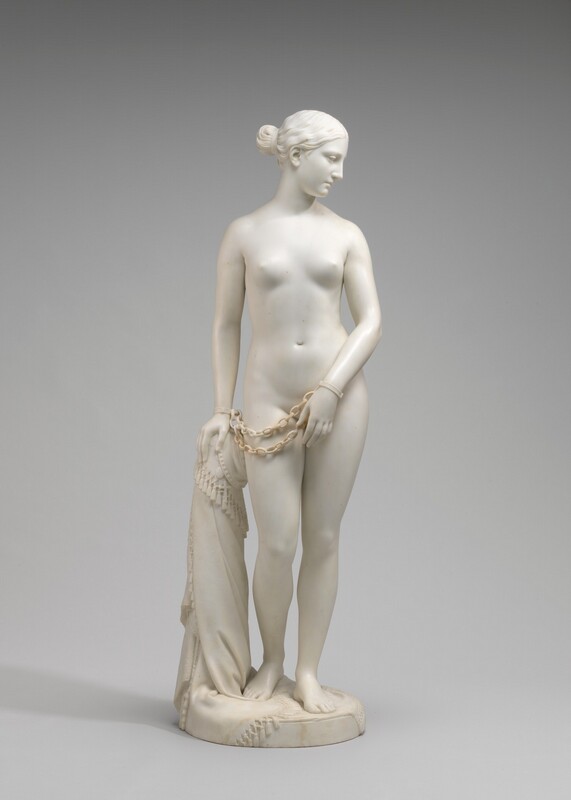 Carved from creamy white marble, a nude woman stands next to a hip-high support, perhaps a low post. In this photograph, her body faces us, and she looks down to our right in profile. Her wavy hair is tucked behind her ear and drawn back in a bun at the nape of her neck. Her weight rests on her left leg, on our right, and her other knee is bent. Her left arm is angled in front of her body so her hand covers her groin. Her other hand, on our left, rests on the post. Chains hang from shackles encircling her wrists. The post is covered with a cloth that gathers around the top and spirals to the ground beneath her feet, the edge trimmed with tassels. A cross and medallion peek out from under the cloth near her hand. She stands on a circular base.