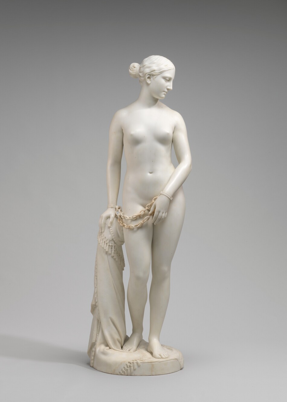 Carved from creamy white marble, a nude woman stands next to a hip-high support, perhaps a low post. In this photograph, her body faces us, and she looks down to our right in profile. Her wavy hair is tucked behind her ear and drawn back in a bun at the nape of her neck. Her weight rests on her left leg, on our right, and her other knee is bent. Her left arm is angled in front of her body so her hand covers her groin. Her other hand, on our left, rests on the post. Chains hang from shackles encircling her wrists. The post is covered with a cloth that gathers around the top and spirals to the ground beneath her feet, the edge trimmed with tassels. A cross and medallion peek out from under the cloth near her hand. She stands on a circular base.