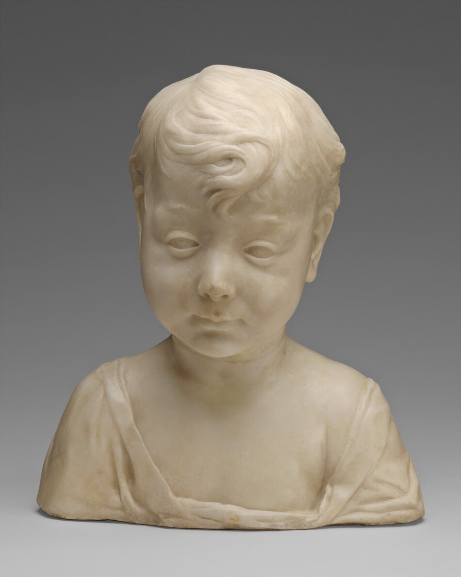 The sculpture depicts the bust of a young child with heavily lidded eyes, thin lips, and chubby cheeks, their face in a neutral expression. The child has short, wavy hair, one prominent curl falling onto their forehead. The child's gaze is directed towards us. They are wearing a draping garment with a wide neckline. The sculpture is made of smooth, light-colored material.