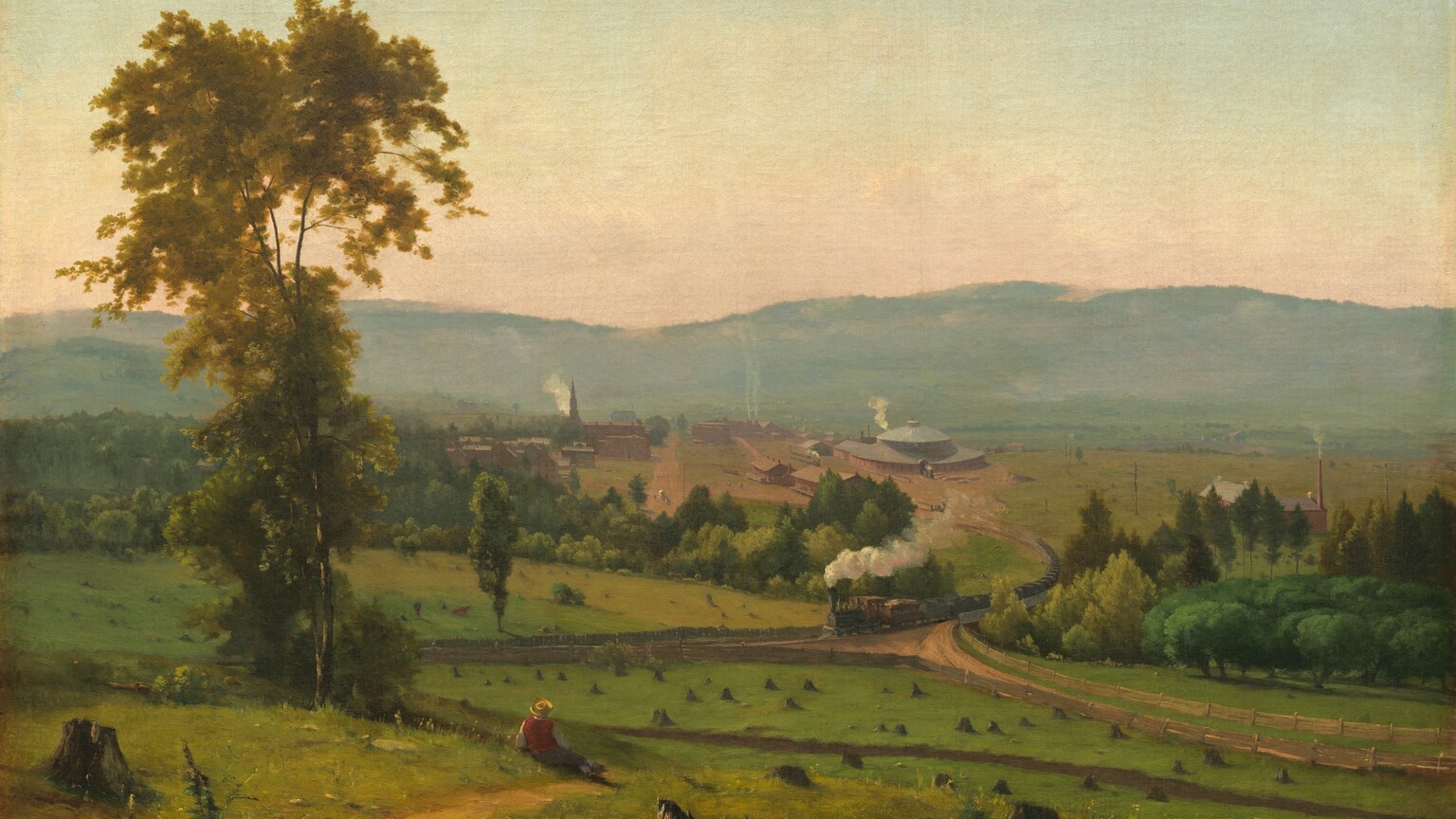 We look across and down into a valley with a person sitting near a tall tree and a train puffing smoke beyond, all enclosed by a band of mountains in the distance in this horizontal landscape painting. Closest to us, several broken, jagged tree stumps are spaced across the painting’s width. A little distance away and to our left, the person wears a yellow, broad-brimmed hat, red vest, and gray pants. He reclines propped on his left elbow near a walking path beside a tall, slender tree with golden leaves. The green meadow stretching in front of him is dotted with tree stumps cut close to the ground. Beyond the meadow, puffs of white smoke trail behind a long steam locomotive that crosses a bridge spanning a tree-filled ravine, headed to our left. The ravine creates a diagonal line across the canvas, moving subtly away from us to our left. The train has climbed out of the valley, away from a cluster of brick-red buildings. The most prominent structure is a train roundhouse, a large building with a high, domed roof to the right of the tracks. Smoke rises from chimneys on long, warehouse-like buildings, and a steeple and smaller structures suggest a church and homes to our left. Hazy in the distance, a row of mountains lines the horizon, which comes about halfway up the composition. The sky above deepens from pale, shell pink over the mountains to watery, pale blue above. The artist signed the work in tiny letters in the lower left corner: “G. Inness.”