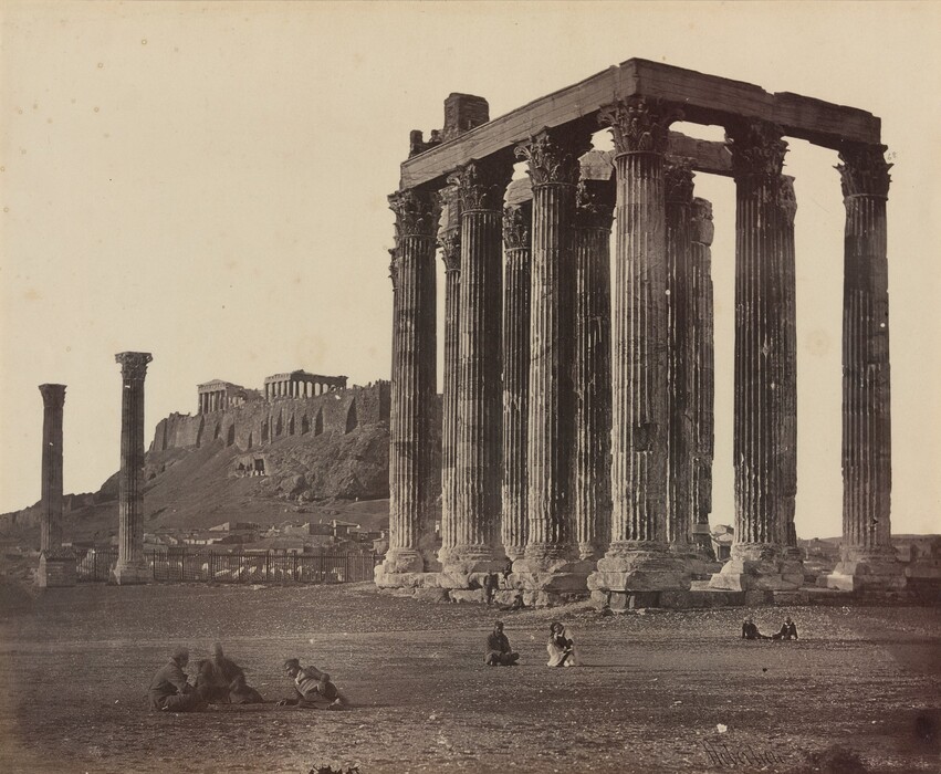The image shows a vintage photograph of an ancient architectural setting with towering columns dominating the view. The perspective is straightforward and at ground level. The horizon is low and distant structures, including a hill with an ancient building complex, are visible. The columns exhibit intricate detailing at their Ionic capitals. The photograph is monochromatic in various shades of sepia. Several figures are seated on the ground, adding a sense of scale.