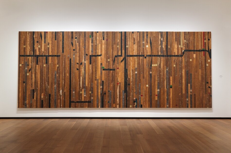 Narrow boards of a wood gymnasium floor were taken apart and reassembled as vertical planks that make up this wide, rectangular panel, which hangs on a wall. Several boards have long strips of black tape and one, near the top right corner, has a band of red tape. Most of the markings are short checks of black, white, red, and green tape. The wood stain is dark in some areas, gray with damage in others, or flaking in places. The two longest horizontal black stripes are near the top left corner and across the right half, about a third of the way down from the top edge of the piece. The room where this hangs has wider planked wood floors and a white wall.