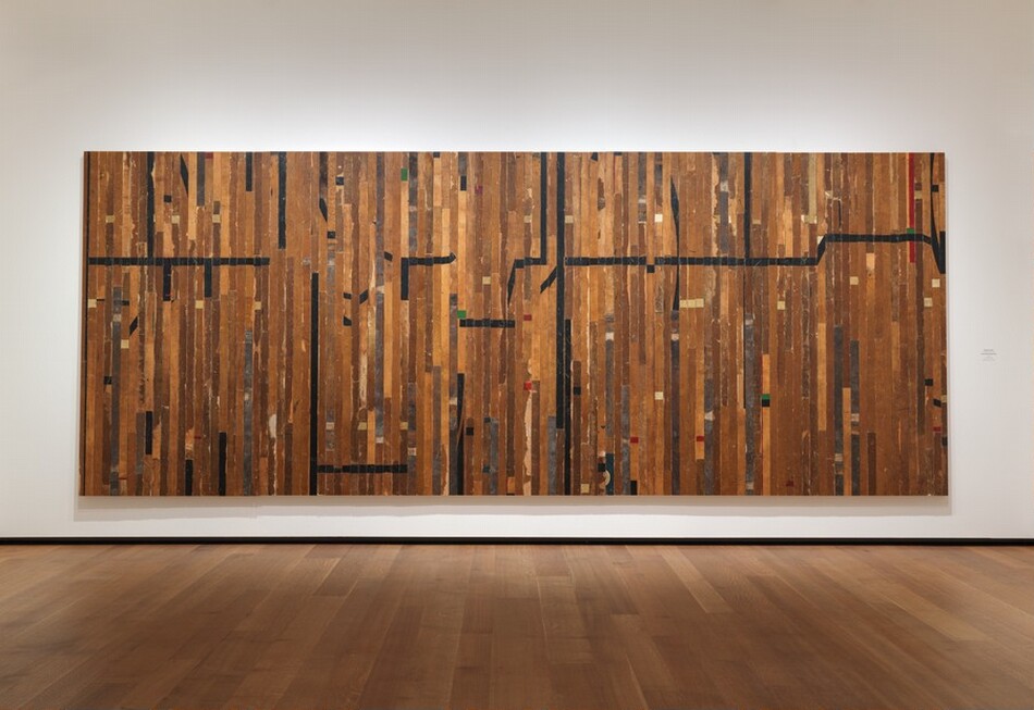 Narrow boards of a wood gymnasium floor were taken apart and reassembled as vertical planks that make up this wide, rectangular panel, which hangs on a wall. Several boards have long strips of black tape and one, near the top right corner, has a band of red tape. Most of the markings are short checks of black, white, red, and green tape. The wood stain is dark in some areas, gray with damage in others, or flaking in places. The two longest horizontal black stripes are near the top left corner and across the right half, about a third of the way down from the top edge of the piece. The room where this hangs has wider planked wood floors and a white wall.
