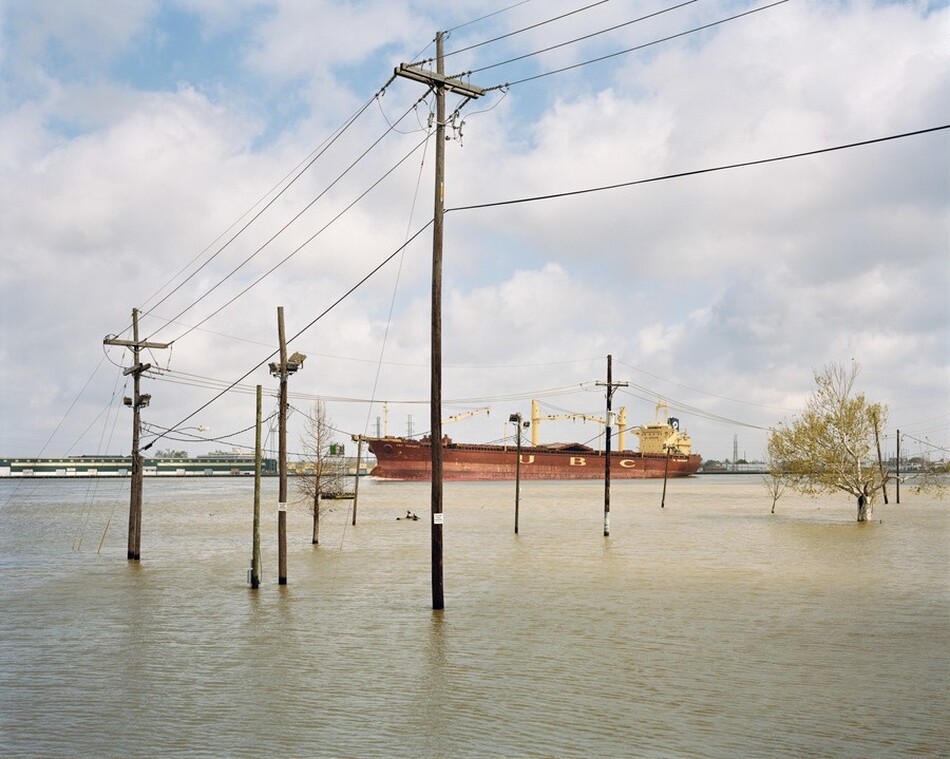 This photograph shows a flooded area with power lines and poles spread out across it, partially submerged in pale brown water. In the distance, a large reddish-brown cargo ship with the letters "U B C" on its rust-red hull is visible. The horizon is in the center of the photograph, where the water meets the sky. The sky is covered with clouds against a light blue background. There are sparse trees submerged in the water. The color palette includes browns, blues, and greens.