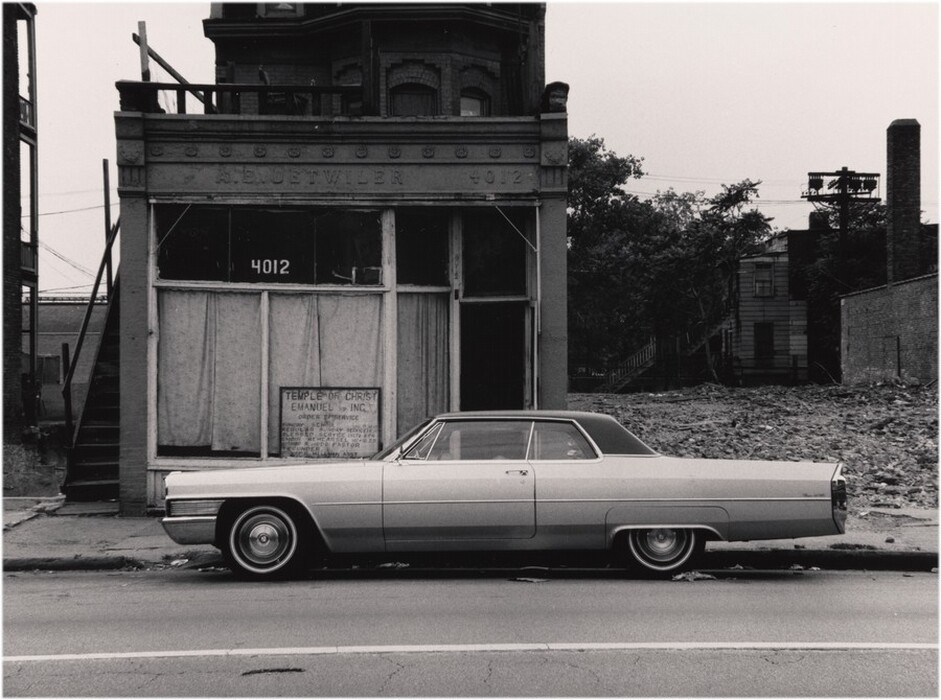 This is a photograph of a vintage automobile parked in front of an old building. The image shows a large, classic car and a weathered building with a sign reading "Temple of Christ Emanuel Inc." The building has boarded-up windows and an upper floor with arched windows, surrounded by a desolate area with rubble and overgrown vegetation. The photograph has a monochrome palette.