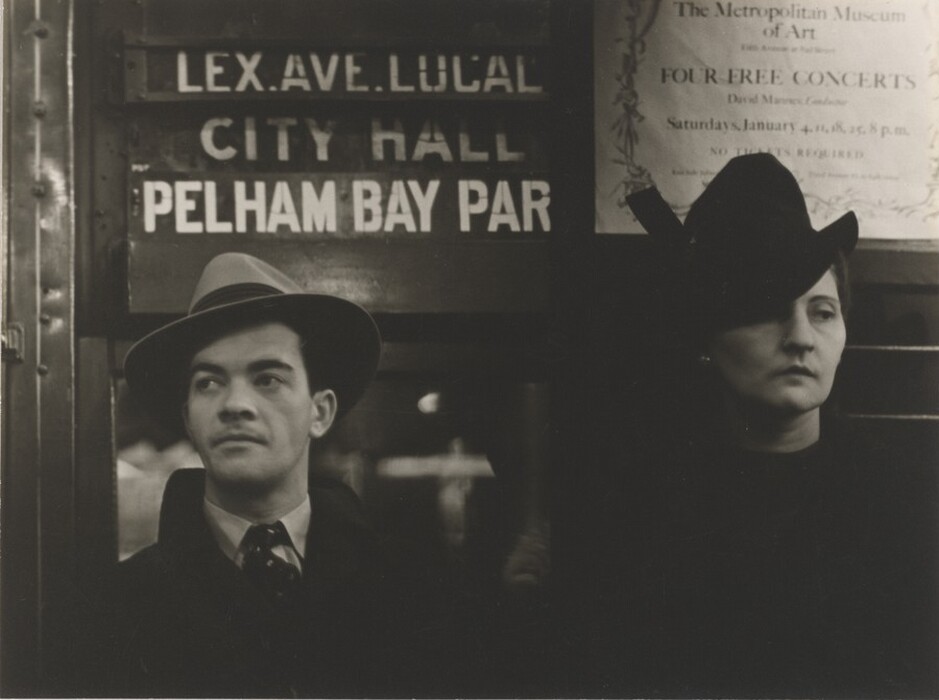 The image displays a partial view of two individuals standing side by side from the waist up. The man is slightly turned to his right, wearing a light-colored fedora hat, dark coat, shirt, and tie. He has dark hair and a focused expression with a strong jawline. The woman is facing forward with light-colored hair under a dark hat, casting a shadow over her eyes. She wears minimal makeup and stud earrings. The background shows subway station signs like "LEX.AVE.LOCAL," "CITY HALL," and "PELHAM BAY PARK," with a poster advertising concerts at "The Metropolitan Museum of Art."