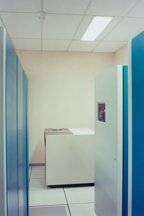 This is a photograph of a computer server room. The image shows a room with light-colored walls and a drop ceiling with fluorescent lighting. The photograph features several large, rectangular server units or computer cabinets. The floors are made of tiles. The room appears clean and organized, with ample space between the equipment, emphasizing a sense of order and functionality typical of server environments.