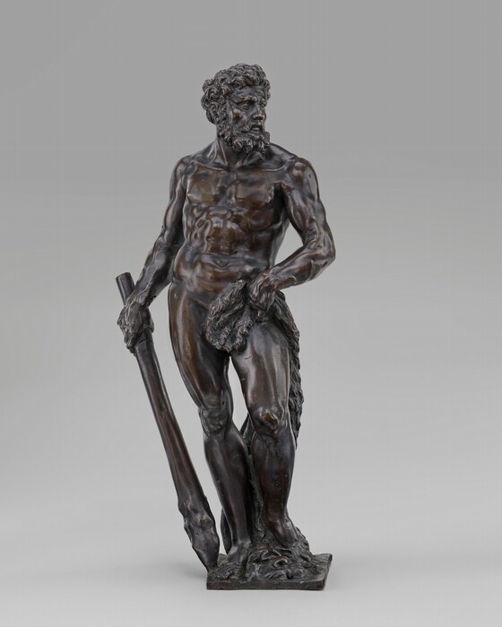The sculpture depicts a bearded, muscular man standing in a contrapposto pose. His left hand grasps a club resting on the ground, with a lion's skin draped over his left shoulder and waist. The material is bronze, displaying a dark metallic sheen and intricate details of the man's hair, beard, and musculature. The life-sized sculpture stands on a square base.