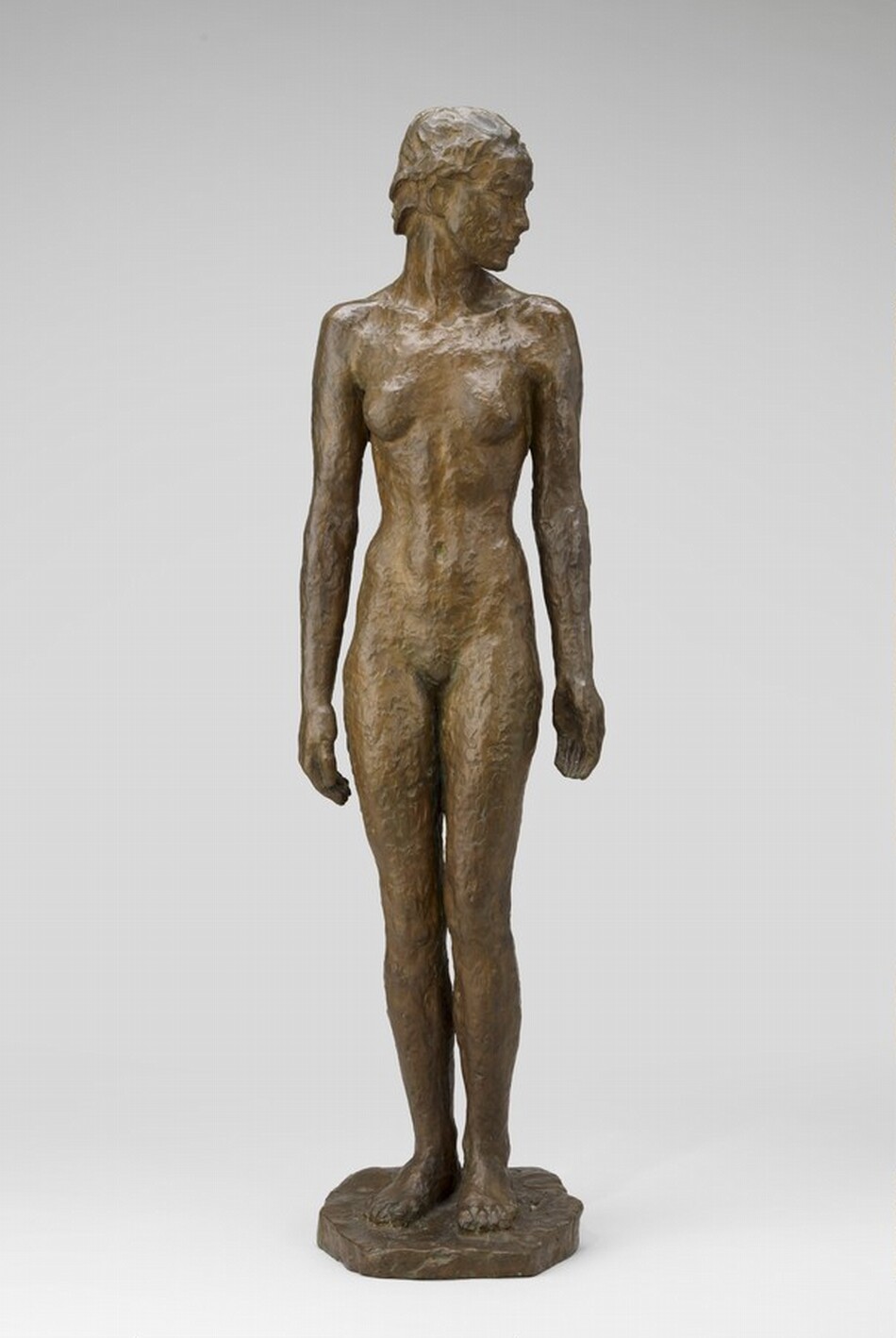 The sculpture is of a standing nude female figure. The subject is depicted in a naturalistic pose with arms down at her sides and her head turned slightly to the right. The sculpture is made from a bronze material, giving it a metallic brown appearance with a textured surface that conveys the form and contours of the human body. The figure stands on a simple base that appears to be part of the same material as the rest of the sculpture.
