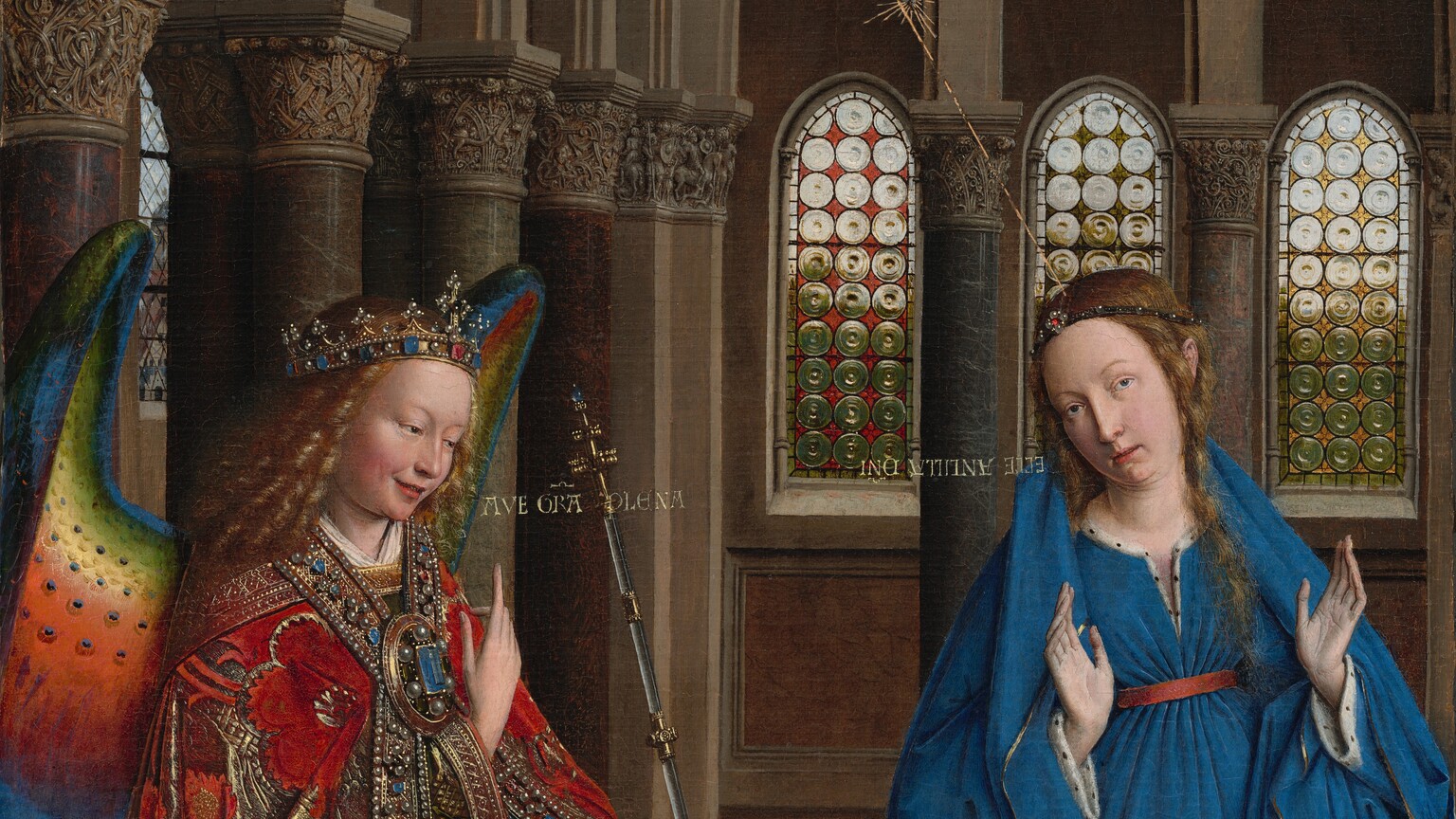 A woman and winged angel, both with pale, peach skin, are situated in a church interior in this tall, narrow painting. To our left, the angel has long, blond, curly hair, smooth skin, and is smiling. The wings are outlined in royal blue, and they blend from blue to green to yellow to crimson. The angel holds one hand, closer to us, up at chest height with the index finger subtly pointing upward. Holding a long scepter in the other hand, the angel angles their body toward the woman to our right. The angel wears a gold jewel and pearl-encrusted crown and a jeweled long, voluminous robe in scarlet-red and shimmering gold brocade. The neck and along the opening down the front are lined with pearls and jewels. The angel looks toward the woman, who wears a royal-blue dress tied with a red belt at the high waist. Her long brown hair is tied back but one tendril falls over her left shoulder, to our right. She kneels facing us with her raised hands facing outward. Her head is tipped a bit to our left, and she looks up and into the distance to our right with lips slightly parted. She kneels behind a book lying open on a low table. A vase of white lilies and a red cushion lies on the floor in front of the table, close to us. The floor is decorated with people and scenes outlined in black and set into square panels, as if inlaid with wood. The church behind and above the people has a row of tall, narrow arches with bull’s-eye glass windows. A walkway lined with columns runs above the arches, and sunlight comes in through arched windows under the flat wood ceiling. A white dove flies toward the woman on gold lines from a window at the upper left of the painting. Latin words painted in gold capital letters are exchanged between the people. The angel says, “AVE GRA PLENA.” The letters of the woman’s response are painted upside down and backward: “ECCE ANCILLA DNI.”