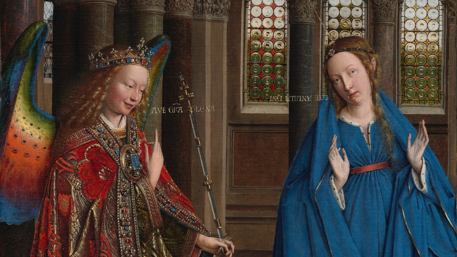 A woman and winged angel, both with pale, peach skin, are situated in a church interior in this tall, narrow painting. To our left, the angel has long, blond, curly hair, smooth skin, and is smiling. The wings are outlined in royal blue, and they blend from blue to green to yellow to crimson. The angel holds one hand, closer to us, up at chest height with the index finger subtly pointing upward. Holding a long scepter in the other hand, the angel angles their body toward the woman to our right. The angel wears a gold jewel and pearl-encrusted crown and a jeweled long, voluminous robe in scarlet-red and shimmering gold brocade. The neck and along the opening down the front are lined with pearls and jewels. The angel looks toward the woman, who wears a royal-blue dress tied with a red belt at the high waist. Her long brown hair is tied back but one tendril falls over her left shoulder, to our right. She kneels facing us with her raised hands facing outward. Her head is tipped a bit to our left, and she looks up and into the distance to our right with lips slightly parted. She kneels behind a book lying open on a low table. A vase of white lilies and a red cushion lies on the floor in front of the table, close to us. The floor is decorated with people and scenes outlined in black and set into square panels, as if inlaid with wood. The church behind and above the people has a row of tall, narrow arches with bull’s-eye glass windows. A walkway lined with columns runs above the arches, and sunlight comes in through arched windows under the flat wood ceiling. A white dove flies toward the woman on gold lines from a window at the upper left of the painting. Latin words painted in gold capital letters are exchanged between the people. The angel says, “AVE GRA PLENA.” The letters of the woman’s response are painted upside down and backward: “ECCE ANCILLA DNI.”
