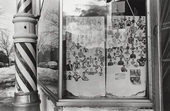 This is an image of a barber shop window. The foreground displays a classic barber's pole with red and white stripes. Inside the window, there is a collage of cut-out photographs featuring muscular individuals in different poses. The two panels that hold the images show signs of weathering at the bottom. Outside, the window reflects a snowy street with cars and leafless trees, suggesting a winter scene.