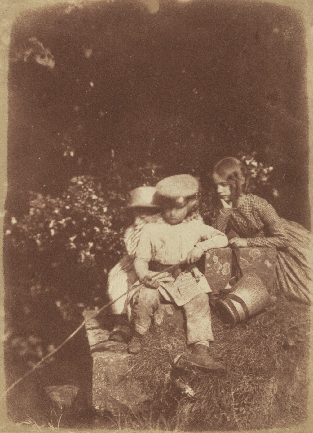 The image shows three children outdoors. One boy is sitting on a rock, holding a stick. Another child is partially obscured behind him. A girl is leaning on the rocks, wearing a dress. A basket is on the grass beside the rock. The background is blurred with dark foliage or trees.