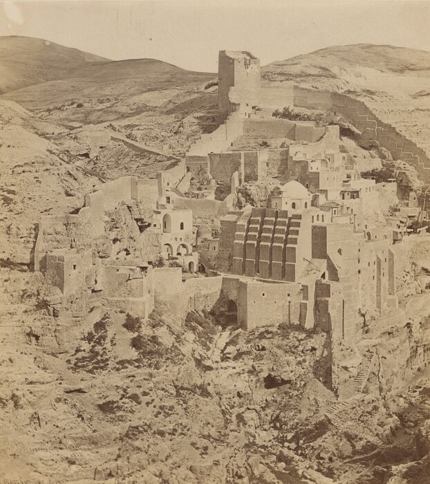 The image shows a stone monastery complex on a mountainside. The buildings are integrated into the natural rock formations, with various terraced structures. The stone walls mimic the color of the landscape, creating a harmonious palette of browns and beiges. The texture of the stone is smooth and varied. The color scheme is monochromatic, adding to the ancient atmosphere of the scene.
