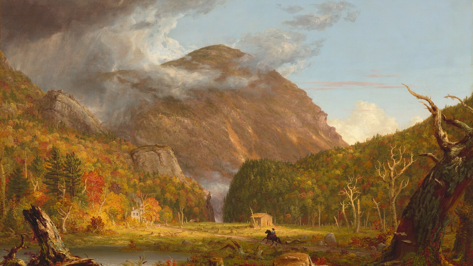 We look across a wide, low clearing at a mountain rising beyond rocky hills, which are mostly covered in pines and blazing red and orange autumn trees, in this horizontal landscape painting. The green grass of the clearing is littered with dead trees and stumps. Beyond a narrow opening between the hills, almost at the center of the composition, a massive mountain towers against the powder-blue sky, nearly reaching the top edge of the painting. The mountain’s jagged face is mottled with warm tones of cinnamon, rust, and caramel brown. The scene is bathed in warm light coming from our right but pewter-gray storm clouds move in from the upper left to encircle the mountain peak. Closest to us, a line of broken tree trunks spans the width of the painting, mixed with scarlet-red, marigold-orange, and green vegetation and reeds. A silvery pond lies just beyond them to our left. Tiny in scale within the landscape, a horse and rider gallop along a wide dirt road emerging from the lower right toward a white house nestled at the foot of the hill to our left. The rider wears a knee-length, denim-blue tunic and red leggings. Upon closer inspection, two children and a white dog become visible, walking toward the rider. A straw-yellow structure sits against the hill to our right. A horse-drawn carriage moves away from the house in the far distance, where the road curves toward the notch. The artist signed and dated the painting as if he wrote directly onto a broken trunk in the lower left corner of the painting: “T. Cole 1839.”