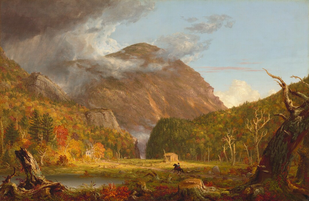 We look across a wide, low clearing at a mountain rising beyond rocky hills, which are mostly covered in pines and blazing red and orange autumn trees, in this horizontal landscape painting. The green grass of the clearing is littered with dead trees and stumps. Beyond a narrow opening between the hills, almost at the center of the composition, a massive mountain towers against the powder-blue sky, nearly reaching the top edge of the painting. The mountain’s jagged face is mottled with warm tones of cinnamon, rust, and caramel brown. The scene is bathed in warm light coming from our right but pewter-gray storm clouds move in from the upper left to encircle the mountain peak. Closest to us, a line of broken tree trunks spans the width of the painting, mixed with scarlet-red, marigold-orange, and green vegetation and reeds. A silvery pond lies just beyond them to our left. Tiny in scale within the landscape, a horse and rider gallop along a wide dirt road emerging from the lower right toward a white house nestled at the foot of the hill to our left. The rider wears a knee-length, denim-blue tunic and red leggings. Upon closer inspection, two children and a white dog become visible, walking toward the rider. A straw-yellow structure sits against the hill to our right. A horse-drawn carriage moves away from the house in the far distance, where the road curves toward the notch. The artist signed and dated the painting as if he wrote directly onto a broken trunk in the lower left corner of the painting: “T. Cole 1839.”