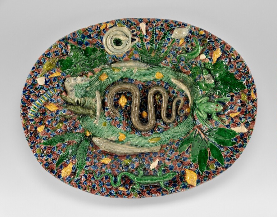 The sculpture is an oval platter featuring detailed elements of creatures and foliage from a natural environment. At its center, there is a coiled snake made of a shiny material, possibly ceramic or glazed pottery. The color palette includes greens, browns, and yellows with accents of blue and white. The snake is surrounded by animals like a frog and fish, as well as plants and leaves. The textures and raised elements create a rich and three-dimensional effect.