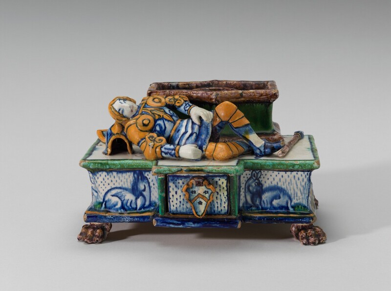 An armored knight and the rectangular trough he lies next to are both on a slightly larger pedestal base in this freestanding, vibrantly colored maiolica inkstand.  The earthenware object was covered with tin-oxide glaze, which, when fired, results in royal blue, grass green, pale orange, and brown hues. The knight’s skin is the white of the earthenware, and his armor is blue and orange. He lies down, using his helmet as a pillow to our left. Both knees are bent, and his hands are by his sides. A staff or lance lies at his feet to our right.  The base has a coat of arms on the center front, and two animals, perhaps deer, lie to either side. The base is outlined in green or blue, and it rests on four brown, paw-shaped feet.