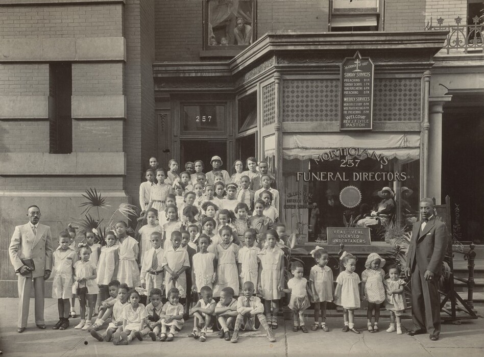 This black-and-white photograph shows a group of people standing and sitting in front of the doorway of a building. The group consists of adults and children of different ages. The group is large, and the people in the back appear to stand on stairs in front of the doorway, while the people in the front stand and sit on the gray sidewalk. The adults are mostly in the back of the group while those in the front are children, although two adult men are positioned on either side of the front row. The man on the left holds a book and wears a light gray suit with a light gray tie, while the man on the right wears a black suit with a white shirt and a black bow tie. Everyone else in the group is dressed in light-colored clothing, with the women and girls wearing dresses and the men and boys wearing collared shirts, some with ties. The people are gathered closely together, most looking at the camera with neutral expressions. The people have a variety of skin tones, from light to medium-toned to dark. Most of them seem to have dark hair. Two buildings frame the group on either side; on the left is an unmarked brick building, and on the right is a building with a large window and a sign labeled "Morticians 257 Funeral Directors." along with other signage. Two people can be seen through this window looking out towards us, and more people are looking out from a window at the top of the photograph.