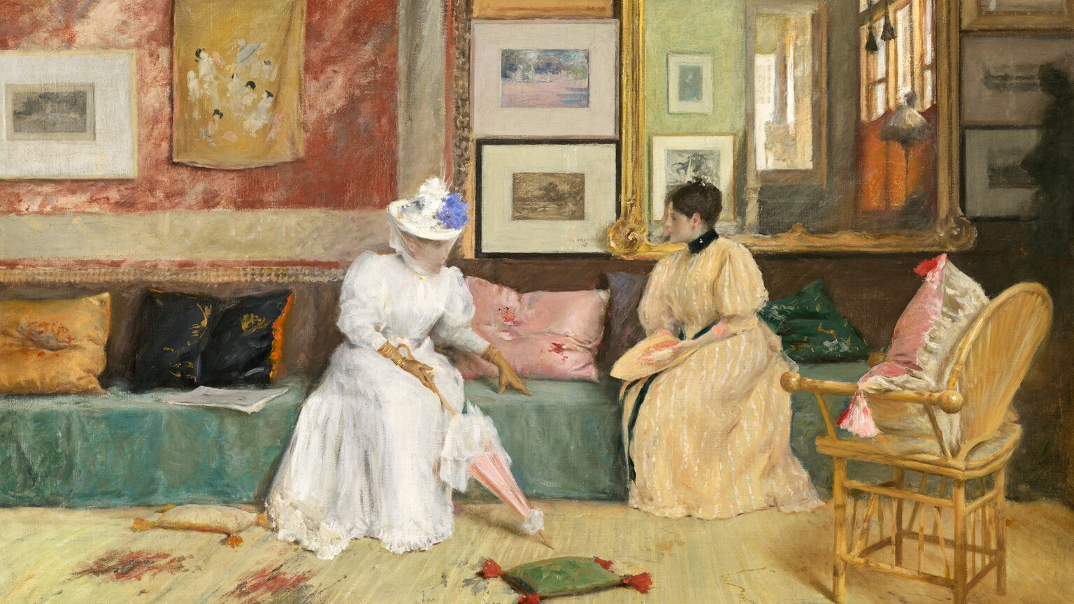 Two pale-skinned women sit facing each other on a long sofa centered on the wall across from us in a room warmly lit from the right in this horizontal painting. The scene is loosely painted so some details are indistinct. The woman on our left wears a white, full-length dress belted at the waist and trimmed with a ruffled hem. The puffy sleeves narrow below the elbow and are tucked into long, fawn-brown gloves. She wears a brimmed hat topped with dabs of white and lapis-blue paint, presumably flowers, with a sheer veil covering her face and chin. Glints of gold at her wrists might be bracelets. Her lower body faces us but her head and torso turn to her left, our right, to face her companion. She leans forward, with her left hand slightly extended, and she rests her fingertips on the cushion between them. Her other hand, closest to us, holds the curving handle of a closed, shell-pink parasol trimmed with white. To our right, the second woman sits with her knees angled to our left as she turns her head in profile, looking at her companion. She wears a floor-length, butter-yellow dress with white, vertical stripes. The dress has a high black collar, and a black ribbon wraps around her waist and falls down the front of the dress. Her sleeves billow at the shoulder and narrow below the elbow. Her dark brown hair is gathered at the top of her head. She holds a flat object in the same butter yellow in her lap that may be a matching hat. The sofa spans almost the entire width of the center of the painting. Its seat is draped in spruce-green fabric, and emerald-green, mauve, rose-pink, black, and gold throw pillows are scattered along its length. The lower half of the wall behind the women is tawny brown while the upper half is divided into four sections. A coral-red and off-white wall hanging fills the wall to our left of the woman in white. A framed artwork and mustard-yellow fabric with daubs of white, ash gray, and black hang on it. Moving right, a column of three framed artworks hang on the wall between the women. Next is a large mirror with a gold frame that hangs behind the woman in yellow. At the far right is another column of three framed artworks. The room behind us is reflected in the large mirror, showing an amber-colored wall with large windows and more framed artworks. Steps leading up to another sunlit room are also visible. A wicker chair with a pink and white pillow sits in the lower right, facing the women at an angle. The floor in front of them fills the lower third of the composition and is covered with a vanilla-yellow carpet streaked with areas of smoke gray and brick red. Two small throw pillows with tassels lie at the feet of the woman in white. The tip of the parasol points to a small sage-green pillow with tomato-red tassels lying on the floor between the women. To our left of the woman in white is a cream-white pillow with marigold-orange tassels. The artist signed and dated the painting in the lower left, “Wm. M. Chase. Copyright 1895.”