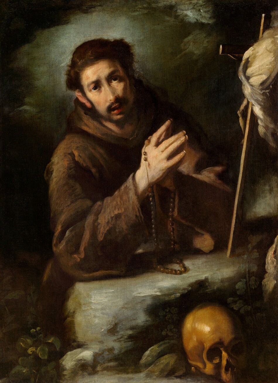 Shown from the waist up, a man with pale, peachy skin and wearing a monk’s robe rests his elbows on a rock with his hands together in prayer in this vertical painting. He is lit from our left, and the background and edges of the composition are mostly in deep shadow. The man’s body and head lean to our left, and he looks at us with dark eyes under gathered brows. He has a long, straight nose, high cheekbones, and his pink lips are parted. He has a dark moustache, and his hair is cut into a ring around his close-cropped crown. He wears a brown hooded robe, and a string of rosary beads hangs from his joined hands. A slender crucifix leaning against a stone in front of him is obscured at the top by what might be a swath of white fabric. The top part of a skull, its surface a golden brown, sits on our side of the rocks. A few plants are tucked in around the skull and boulders. The background is nearly black except for a diffuse pale green halo framing the young man’s head.