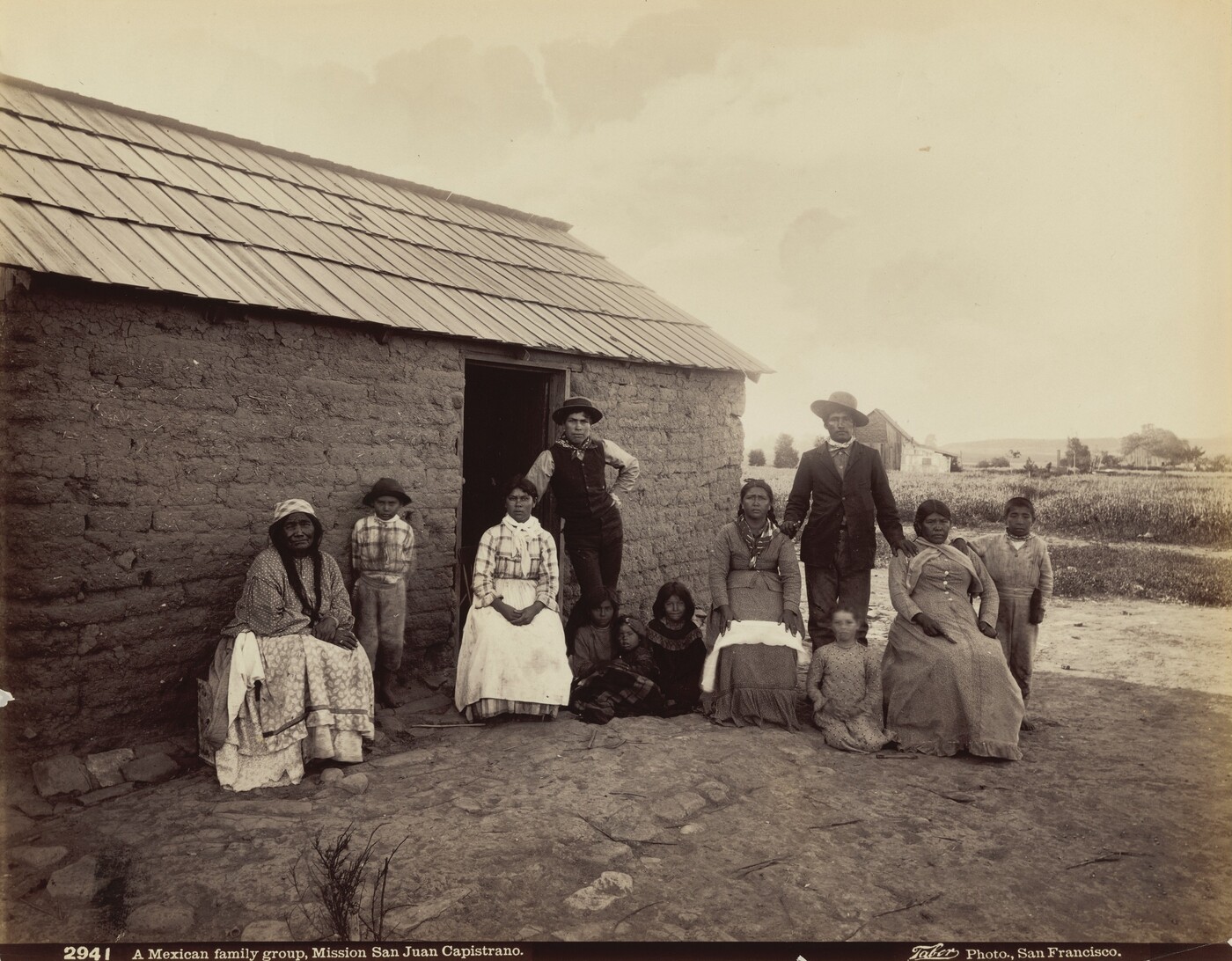 A group of ten people, including men, women, and children, are in front of a rustic adobe house with a shingle roof. The adults are dressed in early 1900s attire, with long skirts and blouses for the women and suits or vests for the men. The children are dressed simply, with some wearing hats. One woman with braids is seated on a wooden bench, and a man in a suit and hat stands behind two seated women. Some children are seated on the ground. Several women wear necklaces. The background shows an open field with sparse vegetation, a few structures, and a cloudy sky.