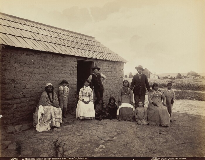 A group of ten people, including men, women, and children, are in front of a rustic adobe house with a shingle roof. The adults are dressed in early 1900s attire, with long skirts and blouses for the women and suits or vests for the men. The children are dressed simply, with some wearing hats. One woman with braids is seated on a wooden bench, and a man in a suit and hat stands behind two seated women. Some children are seated on the ground. Several women wear necklaces. The background shows an open field with sparse vegetation, a few structures, and a cloudy sky.