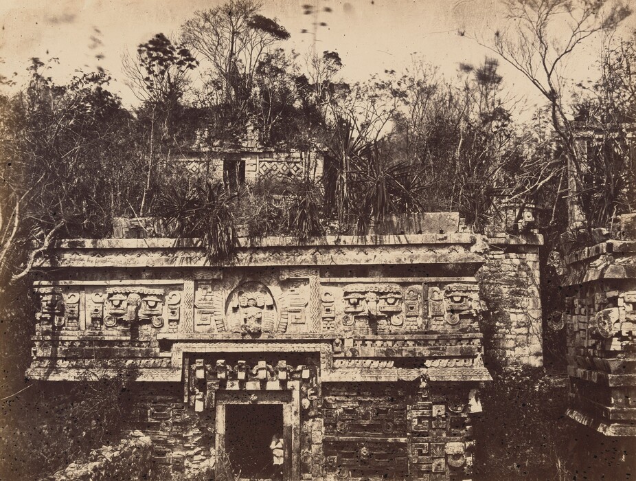 This image shows an ancient stone structure from a frontal low-angle viewpoint. The intricate carvings on the facade are visible, surrounded by dense foliage. Trees in the background create a sense of enclosure, with a sepia-toned color palette emphasizing the weathered textures of the stone. Soft lighting and a vintage quality are highlighted by the soft focus and fine grain of the photograph.