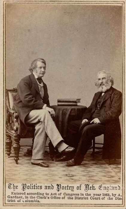 The image shows two men sitting indoors. The man on the left is sitting in a wooden chair, facing the man on the right. He has light-colored hair, is wearing a dark jacket, vest, shirt, necktie, and lighter-colored trousers. The man on the right has a fuller beard and mustache, with medium-length hair, and is wearing a dark three-piece suit. A small table with two books on top is placed between them. The background is plain with a textured surface.