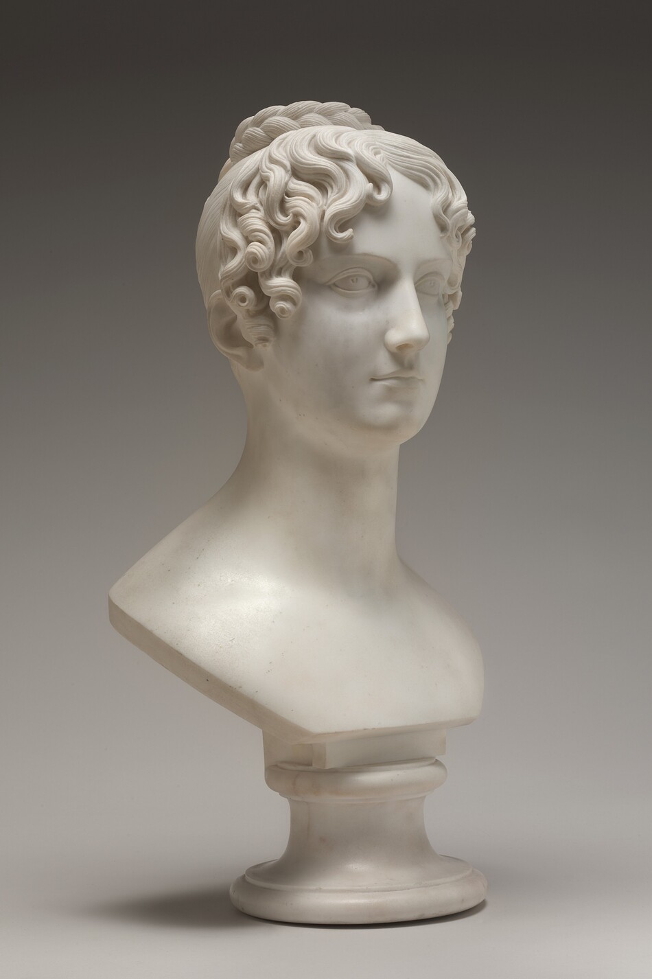 The sculpture is a bust of a person with an elaborate hairstyle. The individual has finely carved, wavy hair gathered neatly at the back. The sculpture is made from white marble. The face of the sculpture displays delicate features, with a straight gaze and a subtle expression. The neck is elongated, and the bust is placed on a simple, rounded pedestal that supports the upper portion of the sculpture.
