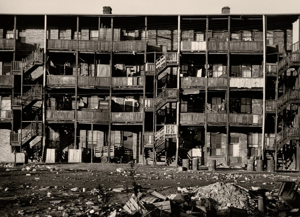 This is a photograph of a row of dilapidated apartment buildings. The image shows a three-story brick structure with wooden balconies on each floor. The balconies appear worn, with some of the wood splintered or broken. There is an assortment of items cluttering the balconies, including barrels, crates, and what may be laundry or fabric hanging from railings. Wooden staircases provide access to the upper floors from the exterior. In the foreground, a lot of debris and litter is scattered across the open ground. The lighting suggests it is daylight, highlighting the texture and condition of the structures.