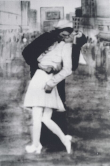The image shows two individuals engaged in an embrace. The man, dressed in a dark naval uniform with a white sailor's cap, leans forward, supporting the woman with one arm around her waist and the other around her shoulders. The woman is wearing a light-colored dress, stockings, and shoes, as she leans back slightly in the man's embrace. She has curly hair partially covered by a nurse's cap, and her head is tilted to one side. The background appears to be an urban setting, with blurred outlines of buildings and a crowd of people.