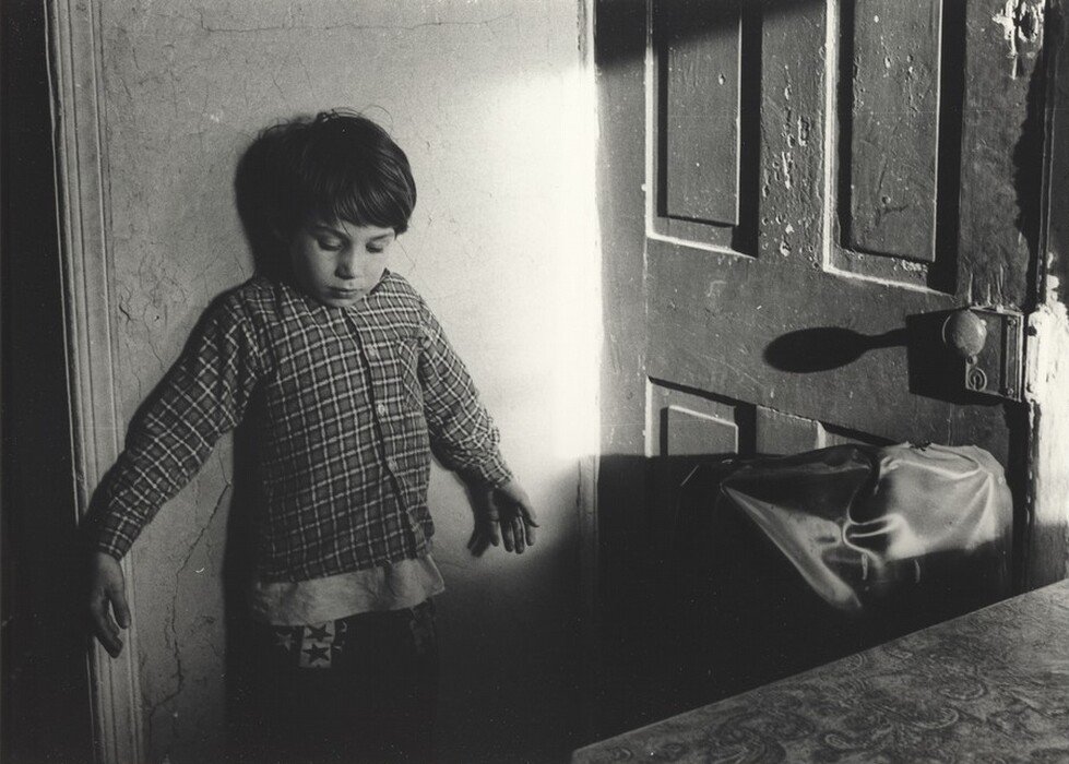 The image shows a young boy from the waist up leaning against a wall. He is positioned with his back to the wall, looking downward. The boy has short, dark hair. He is wearing a patterned plaid shirt with a pocket on the front, and underneath, another shirt or garment is visible, along with pants featuring a star pattern. There is no visible jewelry or objects in his hands. The background consists of a wall with a few visible cracks and a wooden door with peeling paint. Part of a fabric object or bag is propped against the door, and a sunlit table surface is partially visible in the foreground, which appears to have a patterned texture.