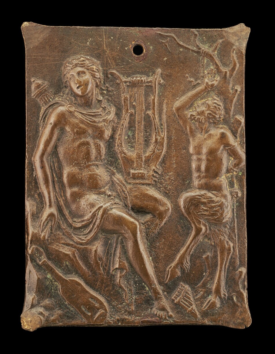The sculpture features two figures in a high-relief style on a rectangular plaque. The figure on the left is depicted clad in draped garments, sitting with one leg crossed over the other, adorned with a laurel wreath on their head, holding a lyre. The figure on the right resembles a satyr, with goat-like legs and animalistic features, posed in a dynamic stance with one arm raised. The sculpture is a metallic bronze hue, indicating metallic material. The plaque shows visible wear and texturing, possibly from age, and includes a circular hole at the top center.