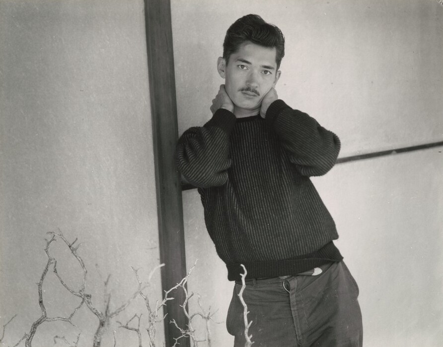 The image depicts a man in a dark sweater with short hair and a mustache. He is leaning against a wooden panel or post with his hands behind his neck, slightly tilted sideways. Dry, leafless branches are visible in the foreground, and the background shows a textured wall with wooden panels.