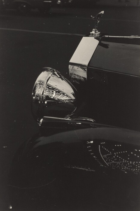 This is a black and white photograph of the front part of a luxury car. The image focuses on the front fender, headlight, and hood ornament of the vehicle. Reflections of lights create patterns on the metal surfaces, emphasizing chrome detailing. The composition is tightly cropped, highlighting the smooth curves and shine of the car's metal parts.