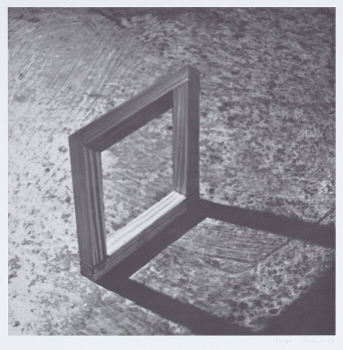 A wooden frame is standing upright on a textured surface, casting a shadow to its side. The image is in grayscale, with shades of gray, black, and white. The frame is in the lower left corner with the shadow extending towards the lower right direction.