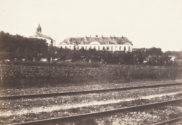 The image displays a large building with a gabled roof and windows in the distance, along with adjacent trees. Nearby is a church-like structure with a steeple partly hidden by trees. In the forefront, there are rows of cultivated plants, potentially a vineyard, divided by wooden fencing. Closest to the viewer are railway tracks running horizontally across the scene. The color scheme consists of sepia and gray tones typical of an old photograph, evoking a vintage and nostalgic atmosphere.
