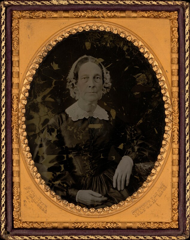 The image shows an older woman sitting composedly, facing the camera. Her facial features appear delicate and wise, with a softly aged complexion. She has a center-parted hairstyle with curls framing her face. She wears a dark gown with a high collar and a white lace overlay. The setting is an ornate, oval-shaped gold frame. The background is dark, keeping the focus on the woman.