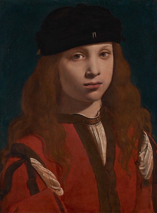 The head, shoulders, and chest of a light-skinned boy or young man wearing a black cap and red garment nearly fill this vertical portrait painting. His body is angled slightly to our right, and he looks at us from the corners of his hazel eyes. He has smooth skin, rounded cheeks, faint, thin eyebrows, and a small nose. His full, dark pink lips are closed. He wears a brimless, soft black cap, slightly askew, over long chestnut-brown hair that covers his ears and flows down past his shoulders. Two small, gold, vertical lines accent the front center of his cap near the lower edge. He wears a collarless, crimson-red jacket, with dark brown trim along the edges and at the shoulder. Pleated folds of his ivory-white shirt material puff out from openings at the shoulders and a V at the top of the chest. There is a also brown band around the neck of the white shirt with a pattern of black, knot-like designs. The white shirt also has two rows of delicate black lines where it is pulled through at the shoulders. The boy is lit from our right and above, casting a shadow on his neck and cheek to our left. The background behind him is teal blue.