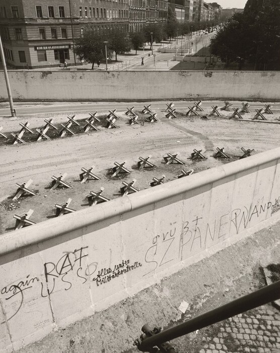 This is a photograph of a concrete barrier with anti-tank obstacles and graffiti. The image shows a fortified barrier with a wide street blocked by anti-tank obstacles. The barrier features a tall concrete wall with various graffiti writings, symbols, and words. Beyond the barrier, there are trees, a sidewalk, and urban buildings in the background. The photo is taken from an elevated view, capturing the surroundings and the tension in the scene.