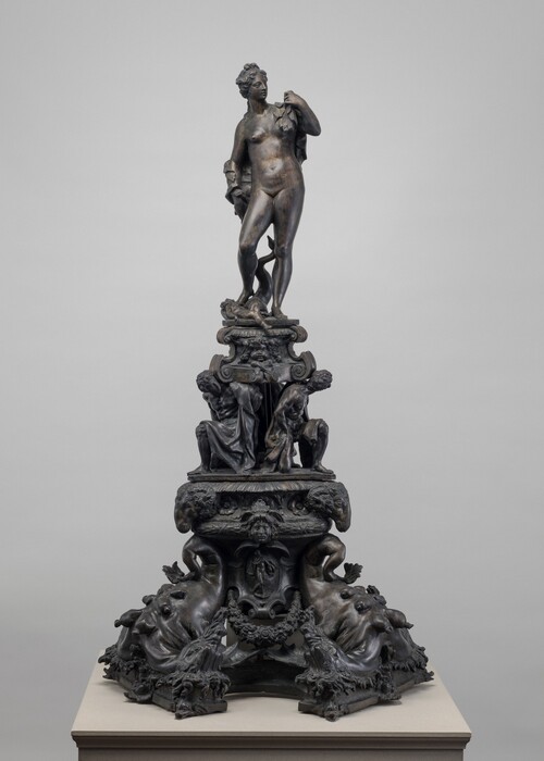 This tall, dark metal sculpture consists of a nude woman standing atop an elaborate, tiered base crowded with human faces, seated people, animal forms, and scrolling ornament. The woman stands with one leg slightly bent and her head turned to our right. Her hair is gathered up, and drapery hangs from her raised left hand and falls behind her shoulder and opposite arm. A twisting, branch-like support rises behind her legs.

Directly below her, two partially draped, curly-haired people sit facing one another beneath a projecting ledge decorated with a central face and curling forms. The broader section below is densely ornamented with additional faces, floral motifs, and animal heads projecting from either side.

At the bottom, four large reclining animal forms, perhaps dogs or other creatures, spread outward to create the sculpture's wide base. Their bodies are covered with rounded protrusions and textured details, and shaggy heads extend toward the corners. Smaller human-like forms and garlands fill the spaces between them. The dark surface varies from nearly black in recessed areas to lighter bronze-gray highlights on projecting forms. The sculpture is photographed against a plain light gray background.