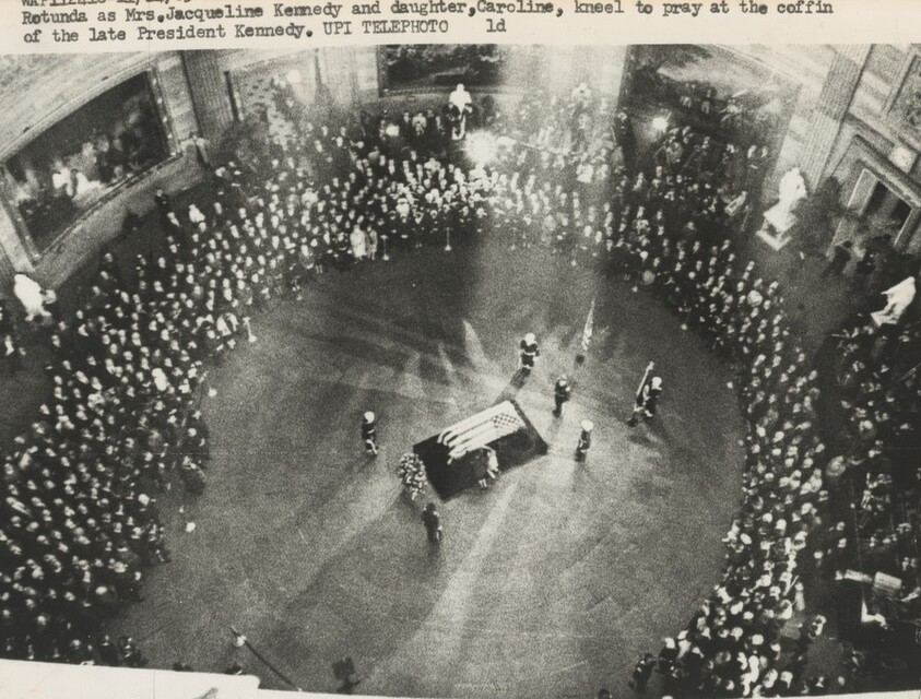 Title from caption on object: “Sun Streams Down on Coffin as Mrs. Kennedy and Caroline Kneel beside It”