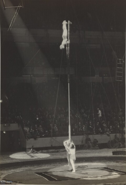 This is a photograph of a circus performance featuring a balancing act. Two performers are engaged in an acrobatic display: one is positioned upside down at the top of a vertical pole while the other supports them from below, holding onto the pole. The scene is set inside a large arena packed with onlookers seated in rows. The photograph captures the performance under a dramatic spotlight, emphasizing the height of the pole and focusing attention on the skill involved in the act. The background reveals an audience with indistinct faces, while the foreground is dominated by the performers and the neatly arranged performance space, which includes apparatuses and a safety mat or platform on the ground. The image has a vintage quality, marked by its monochrome color scheme, typical of early 20th-century photography.