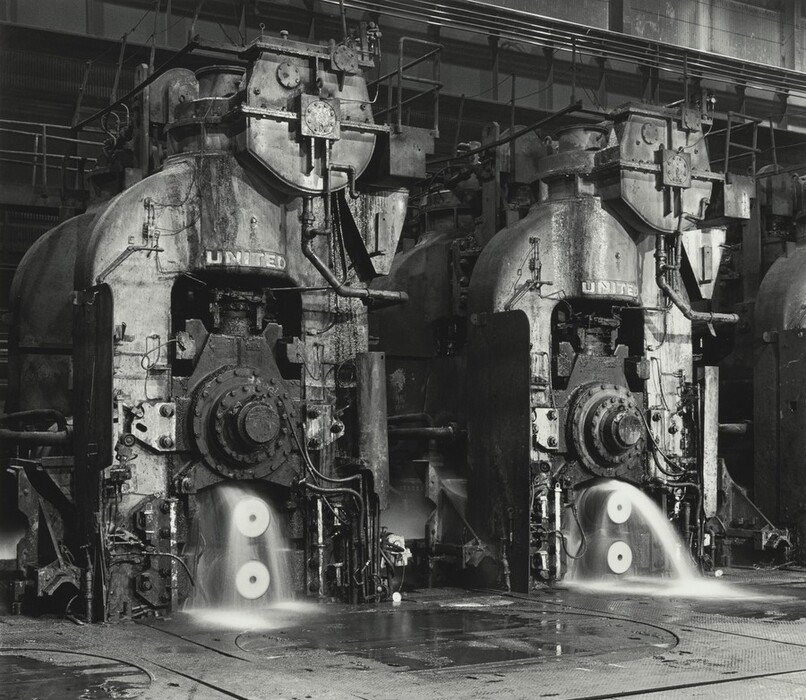 This is a photograph of industrial machinery in a factory setting. The image features two large machines with large gears and hydraulic systems. The machines are labeled with "UNITED." They appear worn with grime and oil stains. Water jets are flowing onto the machinery, potentially for cooling purposes. The image conveys industrial power and mechanical intricacies in manufacturing. The tone of the image is monochromatic.