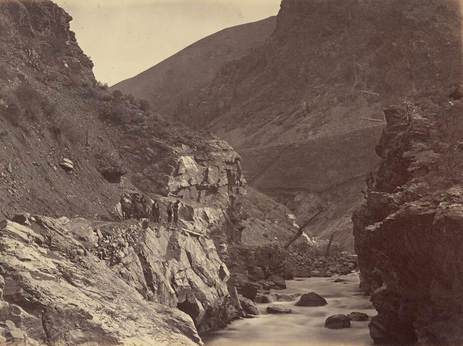 The image shows a rugged mountain landscape with a river flowing from right to left. The view is dominated by the sloping mountain sides and the deep valley carved out by the river. In the foreground, a rocky path winds along the mountainside with several figures and pack animals lined up. The scene is captured in sepia tone, giving it an antique feel. The rocks and flowing water are clear, showing a contrast between the textures. The mountains in the distance gradually fade, adding depth.