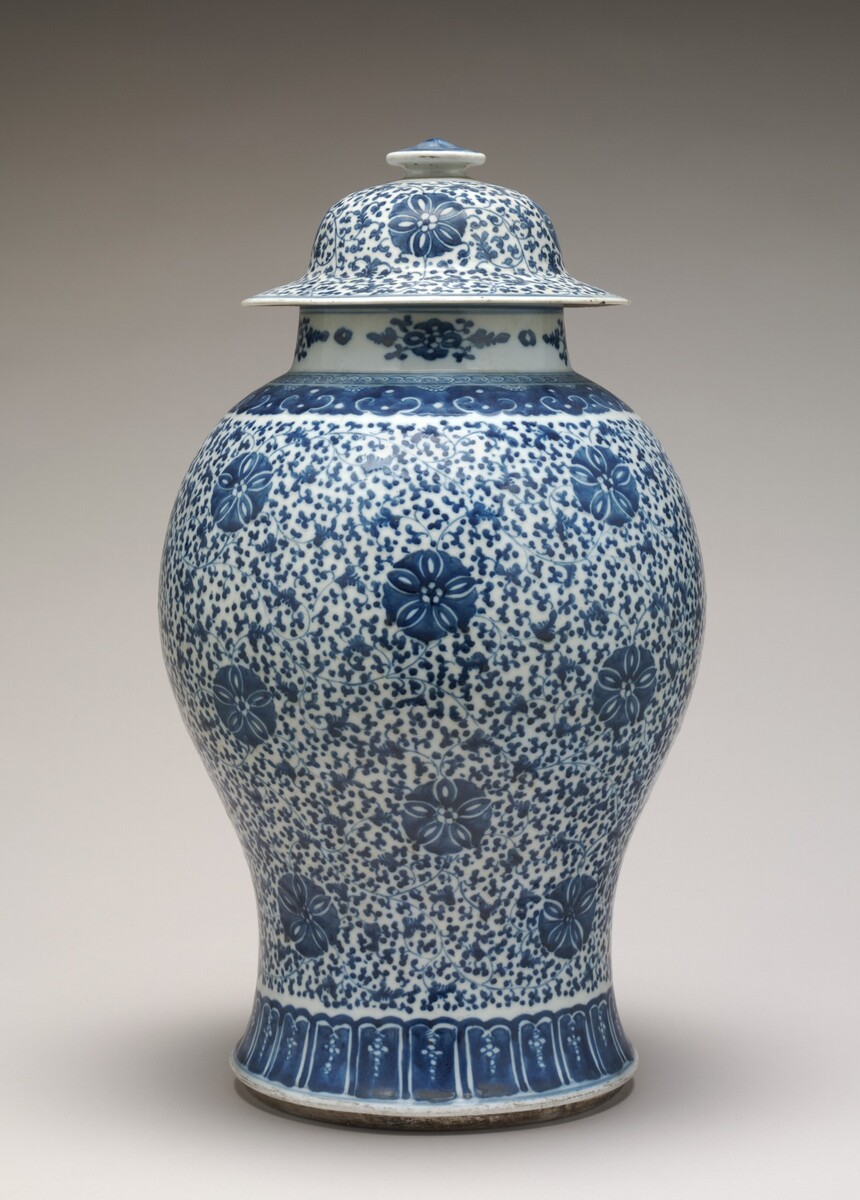This is a photograph of a decorative lidded porcelain vase with blue and white patterns. The vase has an intricate floral design painted in shades of blue against a white background. The body of the vase tapers down to a narrow base and is topped with a fitted lid that has a small finial at the top. The patterns consist of floral motifs interspersed with small dotted designs. The blue used is a deep cobalt shade commonly associated with traditional Chinese porcelain.