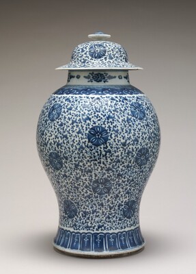 This is a photograph of a decorative lidded porcelain vase with blue and white patterns. The vase has an intricate floral design painted in shades of blue against a white background. The body of the vase tapers down to a narrow base and is topped with a fitted lid that has a small finial at the top. The patterns consist of floral motifs interspersed with small dotted designs. The blue used is a deep cobalt shade commonly associated with traditional Chinese porcelain.
