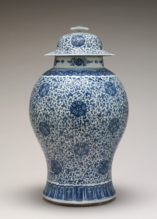 This is a photograph of a decorative lidded porcelain vase with blue and white patterns. The vase has an intricate floral design painted in shades of blue against a white background. The body of the vase tapers down to a narrow base and is topped with a fitted lid that has a small finial at the top. The patterns consist of floral motifs interspersed with small dotted designs. The blue used is a deep cobalt shade commonly associated with traditional Chinese porcelain.