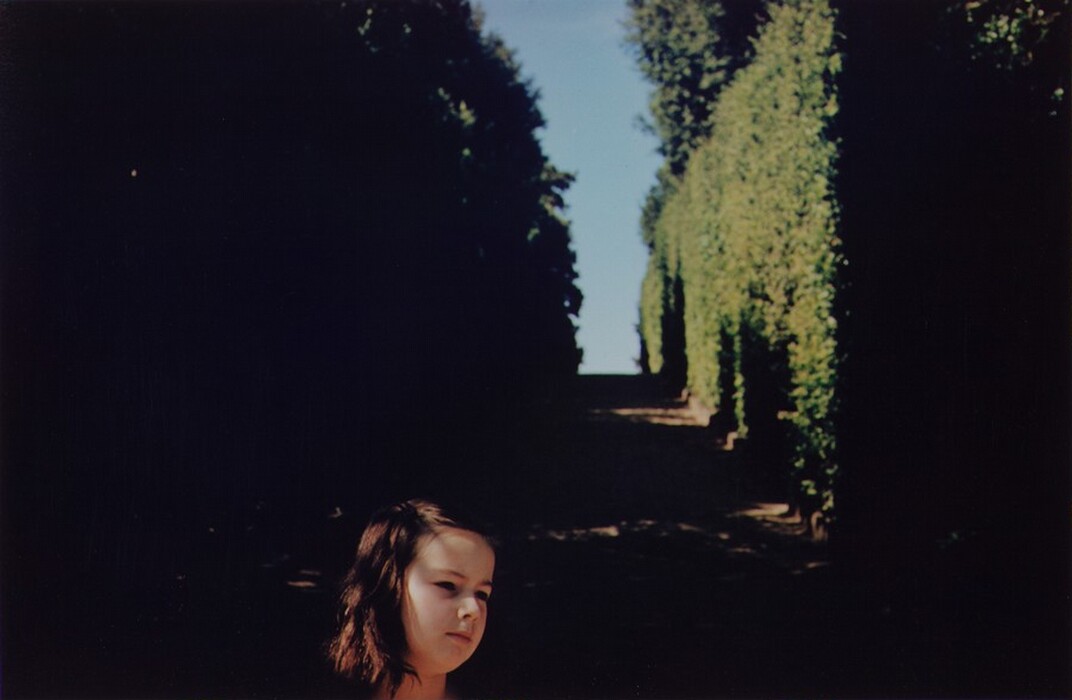 The image shows a young girl's head and shoulders in the lower right corner. She is turned slightly to the left with a neutral expression. She has medium-length dark hair and is wearing clothing out of view. The background shows a hedged pathway with green foliage, shadow, light patches, and a clear blue sky.